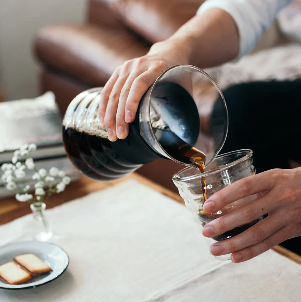 Person pouring coffee from a carafe into a glass on a table with a plate of bread.